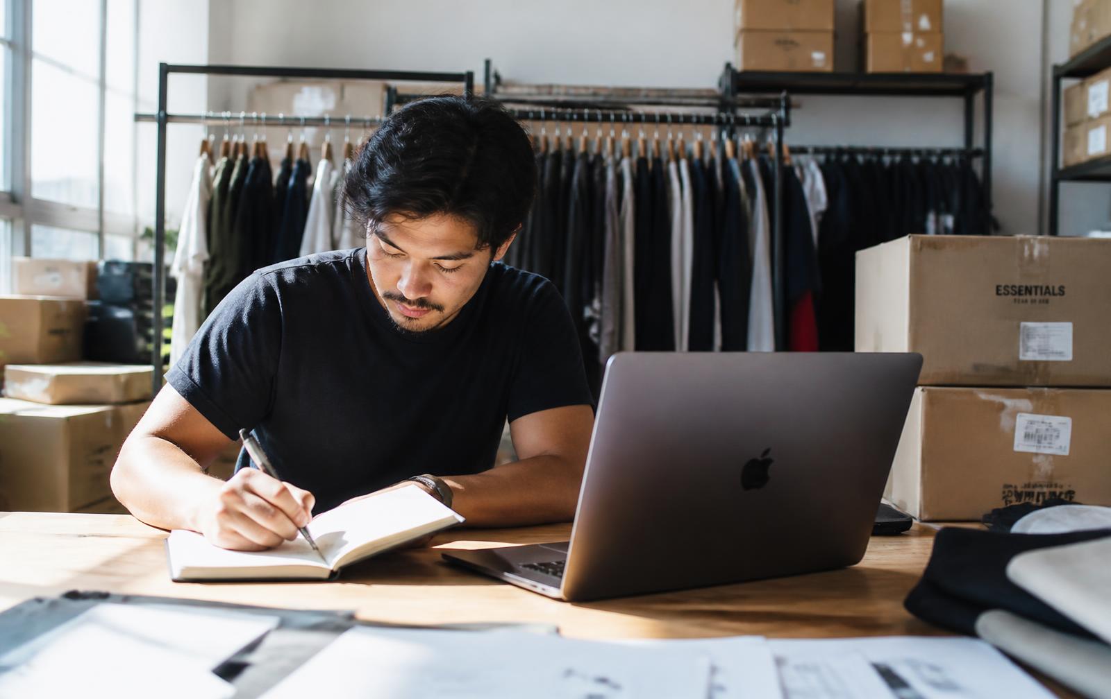 Founder of a small apparel label taking notes beside a laptop in a studio warehouse